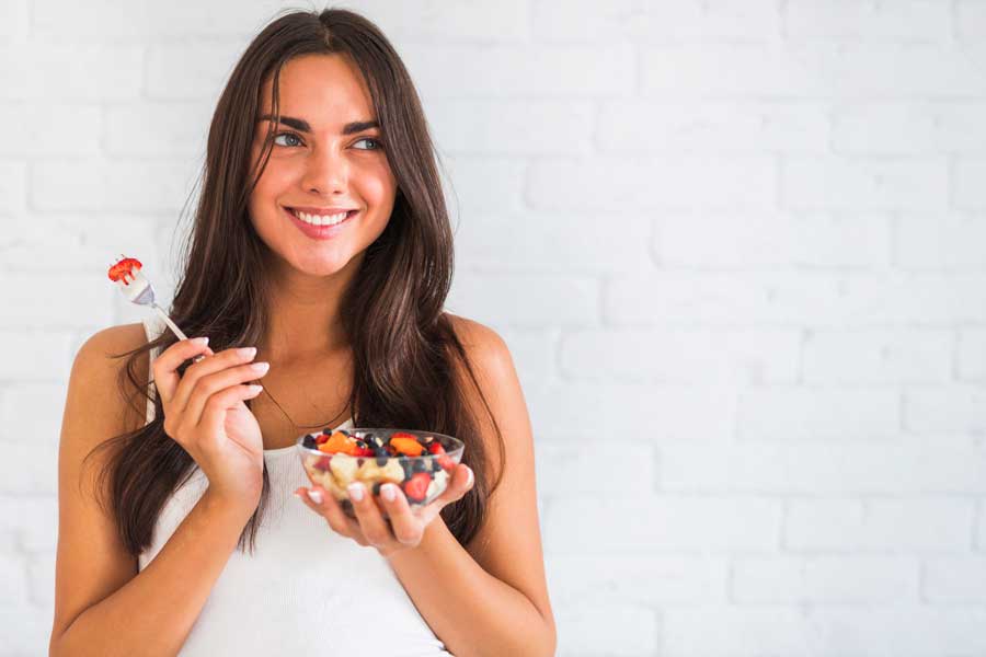 Jovem pensativa sorrindo segurando uma tigela de salada de frutas.